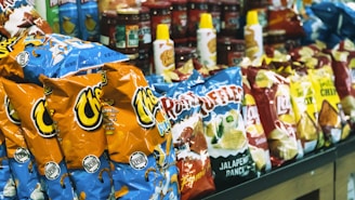 A shelf in a store stocked with a variety of snack products including bags of Cheetos, Ruffles, Lay's chips, and bottles of condiments.