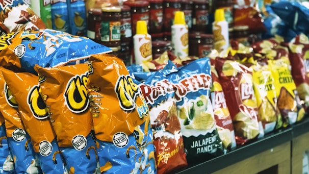 A shelf in a store stocked with a variety of snack products including bags of Cheetos, Ruffles, Lay's chips, and bottles of condiments.