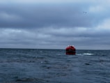 A boat filled with people wearing red life jackets is navigating through a large, grayish sea. The sky above is overcast with dark clouds, suggesting inclement weather. A few birds are flying in the sky.