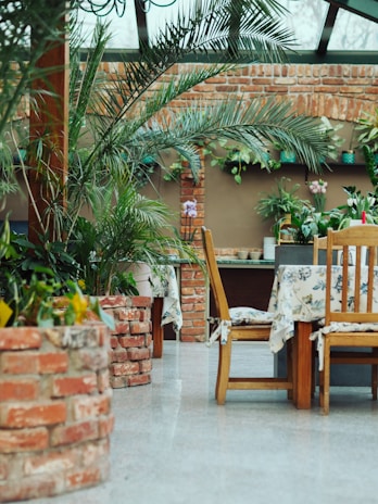 A cozy indoor space surrounded by lush green plants and rustic brick walls. Wooden chairs and tables covered with floral tablecloths create a welcoming atmosphere. The natural light filtering through the ceiling windows adds to the serene ambiance.