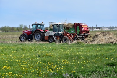 A vibrant agricultural scene with two tractors working on a field surrounded by green grass and scattered yellow flowers. The tractors are red and white with attached machinery, handling hay or straw. In the distant background, there are trees and open fields under a clear blue sky.