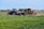 A vibrant agricultural scene with two tractors working on a field surrounded by green grass and scattered yellow flowers. The tractors are red and white with attached machinery, handling hay or straw. In the distant background, there are trees and open fields under a clear blue sky.