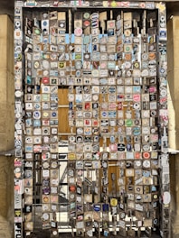 A metal gate is covered with numerous stickers and decals of various shapes, colors, and designs. The background shows a glimpse of a wooden structure or panels, and the eclectic collection of stickers creates a chaotic and vibrant collage.