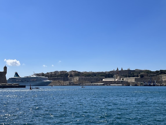 A large cruise ship is docked in a harbor, with a backdrop of historic buildings on the shoreline. The sky is clear and blue, and the water is a deep shade of blue, reflecting the sunlight.