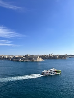 A ferry with bright markings moves across a calm blue sea, leaving a trail of white waves. In the background, there is a historic coastal cityscape with stone buildings under a clear blue sky.