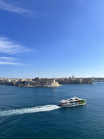 A ferry with bright markings moves across a calm blue sea, leaving a trail of white waves. In the background, there is a historic coastal cityscape with stone buildings under a clear blue sky.