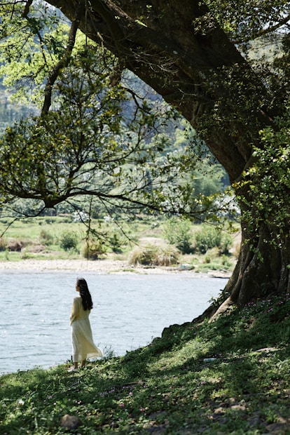 A serene scene featuring a woman in a white dress standing by a tranquil body of water, with lush greenery and a large tree providing shade. The landscape is bathed in soft, natural light, creating a peaceful atmosphere.