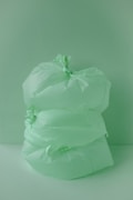 Three large, green plastic bags are stacked against a matching green background, creating a monochromatic scene. The bags are knotted at the top, giving them a full and somewhat rounded appearance.