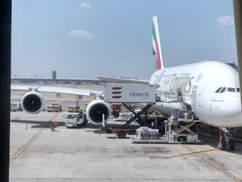 A large passenger airplane is parked at an airport gate with ground support equipment. The aircraft features the logo of an airline on its fuselage. Various cargo containers and vehicles can be seen around, indicating loading or unloading activities. The sky is clear with a few clouds.