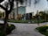 A modern building with reflective glass surfaces stands at the center, surrounded by trees and green grass. A few people are walking on the paved paths leading to the entrance. A sign reads 'Welcome to Mesa Court.' The scene suggests a campus or office park setting.