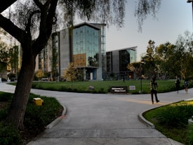 A modern building with reflective glass surfaces stands at the center, surrounded by trees and green grass. A few people are walking on the paved paths leading to the entrance. A sign reads 'Welcome to Mesa Court.' The scene suggests a campus or office park setting.