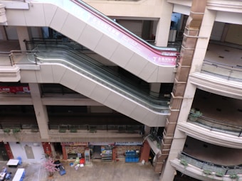 A multi-story indoor space with empty escalators connecting different floors. On the ground level, there are closed or shuttered storefronts, various items scattered including a pink plant, and a couple of small tables with chairs. The area seems deserted, there are no people present in the image.