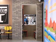 A small kitchen or service area is visible through an open doorway, where a person in a blue uniform and cap is working. The room has a coffee machine and other kitchen equipment. On either side of the doorway, there are dining areas with colorful chairs and wall decorations. One wall features a large poster with vibrant colors and text, while another has a monochrome illustration. There is a brick wall between the dining areas.