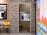 A small kitchen or service area is visible through an open doorway, where a person in a blue uniform and cap is working. The room has a coffee machine and other kitchen equipment. On either side of the doorway, there are dining areas with colorful chairs and wall decorations. One wall features a large poster with vibrant colors and text, while another has a monochrome illustration. There is a brick wall between the dining areas.