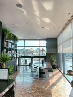 A modern indoor space features large floor-to-ceiling windows allowing natural light to flood the room. The polished marble floor reflects the sunlight, adding warmth to the interior. Along the left side, there are shelves with plants and books, adding a touch of greenery. A long table with a few chairs is positioned centrally, suggesting a dining or meeting area. Various green plants are placed strategically around the room, enhancing the natural aesthetic.