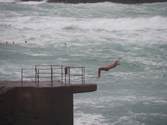 A person wearing swim shorts is captured mid-dive off a concrete platform into rough sea waters. The platform has a metal railing. Several people with swim caps can be seen in the water, indicating that they might be participating in a swimming activity.