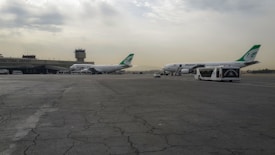 Two large passenger airplanes are stationed on the tarmac near an airport terminal building with visible green tail fins. The runway is cracked and shows signs of wear, while buses and ground vehicles are parked alongside the planes. A control tower stands in the background under a cloudy sky.
