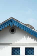 A gable roof with scalloped wooden trim painted blue contrasts against a light blue sky. Below the gable are two square lattice windows. The structure and colors give a quaint and traditional architectural feel.