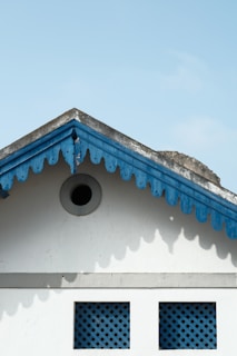 A gable roof with scalloped wooden trim painted blue contrasts against a light blue sky. Below the gable are two square lattice windows. The structure and colors give a quaint and traditional architectural feel.
