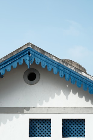 A gable roof with scalloped wooden trim painted blue contrasts against a light blue sky. Below the gable are two square lattice windows. The structure and colors give a quaint and traditional architectural feel.
