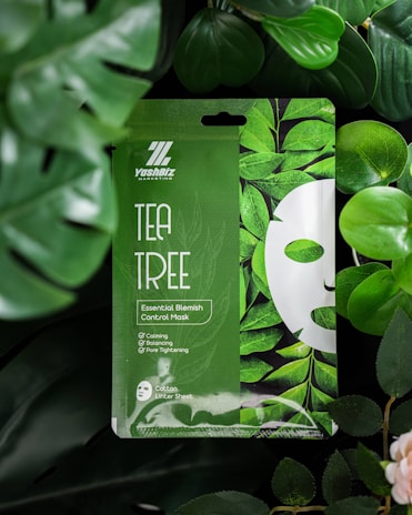 A package of tea tree essential blemish control mask is surrounded by lush green leaves. The packaging is predominantly green with a leaf design and features the words 'Tea Tree' and 'Essential Blemish Control Mask' prominently. It highlights benefits such as calming, balancing, and pore tightening. The environment conveys a natural and refreshing aesthetic.