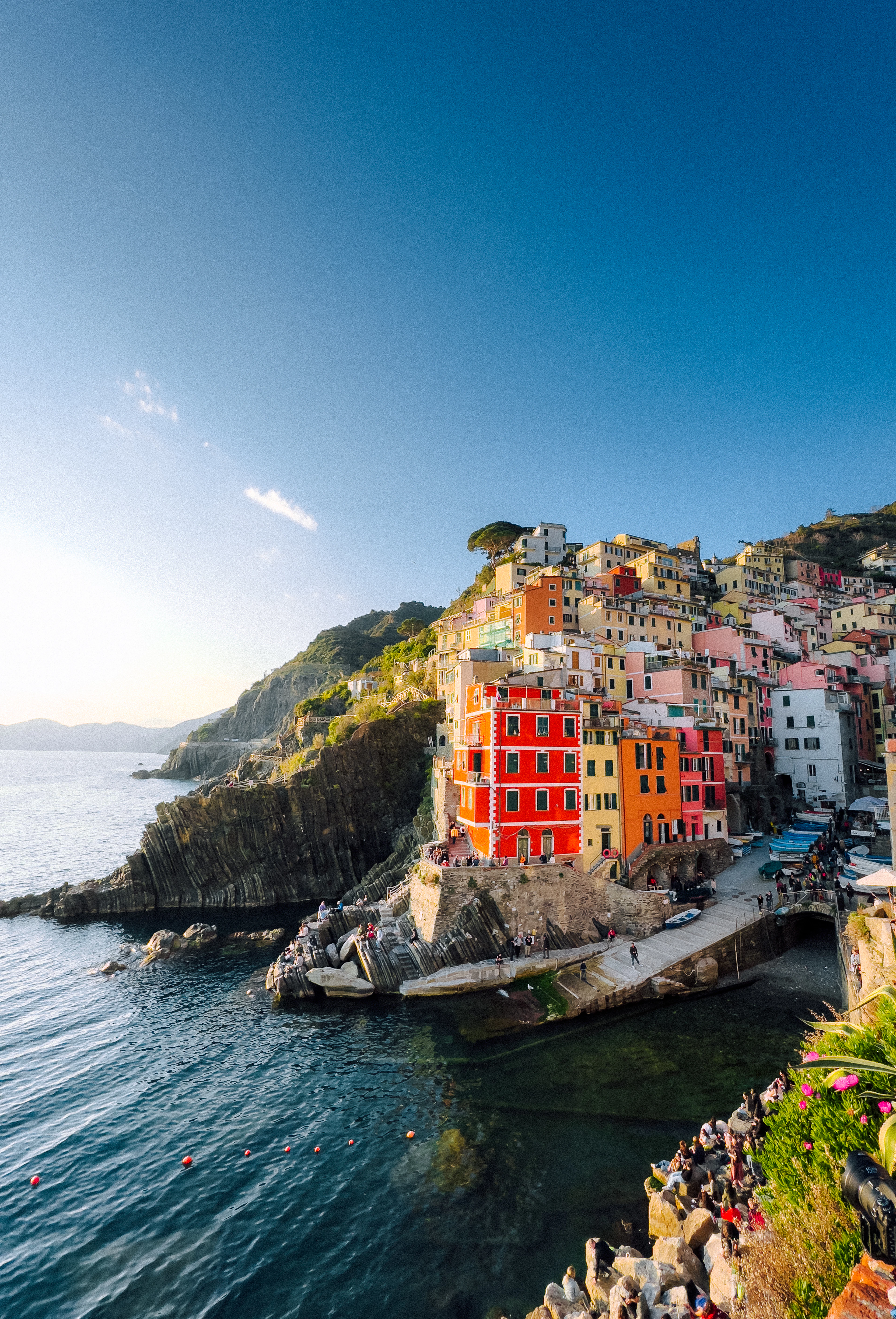 Riomaggiore in late afternoon light, its scarlet and ochre houses stacked above the slipway and small harbour, fishing boats drawn up on the quay and the Ligurian Sea running out to a hazy headland