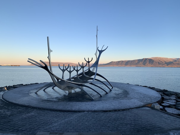 A large metal sculpture resembling a stylized Viking ship is set against a backdrop of a calm body of water and distant mountains under a clear sky. The sculpture is placed on a circular stone platform, creating an artistic and serene scene.