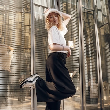 A person poses stylishly in front of a reflective glass wall with horizontal lines. They wear a white shirt, a white bucket hat, black pants, and sneakers. The light plays off the glass, creating an urban, modern atmosphere.