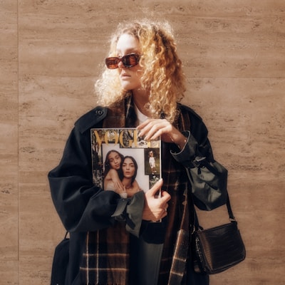 A person with curly blonde hair wearing sunglasses is holding a fashion magazine. The individual is dressed in a dark coat with a scarf and has a black handbag. The background is a textured stone wall, and the sun is casting shadows.