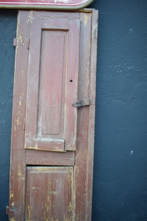 A weathered wooden door with chipped paint stands against a deep blue wall. The top of the door is adorned with a partial red and white sign, suggesting an old establishment. The door's worn texture and the peeling paint add a rustic charm to the scene.
