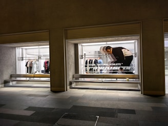 A store display with two large glass windows, featuring clothing racks with various items such as shirts and jackets. There is a poster of a person bending down, with text 'Natural Athlete' across it. The surrounding area includes a tiled walkway.