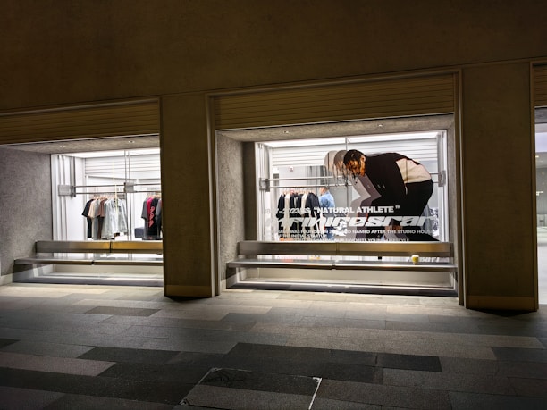A store display with two large glass windows, featuring clothing racks with various items such as shirts and jackets. There is a poster of a person bending down, with text 'Natural Athlete' across it. The surrounding area includes a tiled walkway.
