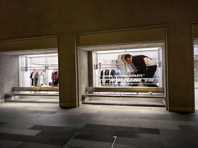 A store display with two large glass windows, featuring clothing racks with various items such as shirts and jackets. There is a poster of a person bending down, with text 'Natural Athlete' across it. The surrounding area includes a tiled walkway.