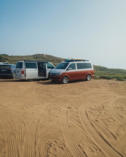 Several vans are parked on a sandy area with clear tire tracks. The setting appears to be a rugged outdoor location with grassy hills in the background. The van doors are open and surfboards are visible on top of one van.