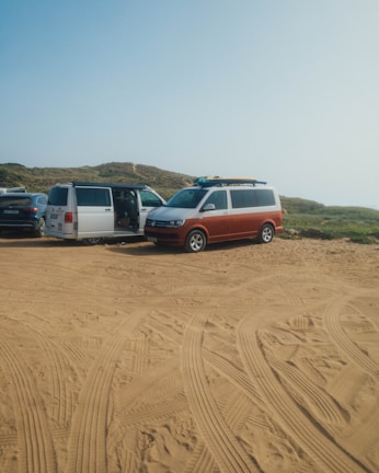 Several vans are parked on a sandy area with clear tire tracks. The setting appears to be a rugged outdoor location with grassy hills in the background. The van doors are open and surfboards are visible on top of one van.