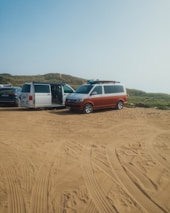 Several vans are parked on a sandy area with clear tire tracks. The setting appears to be a rugged outdoor location with grassy hills in the background. The van doors are open and surfboards are visible on top of one van.