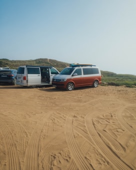 Several vans are parked on a sandy area with clear tire tracks. The setting appears to be a rugged outdoor location with grassy hills in the background. The van doors are open and surfboards are visible on top of one van.
