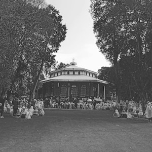 A large group of people, many wearing light-colored clothing, are gathered in front of a historic, round building surrounded by tall trees. The scene is in black and white, with people seated on the ground or standing around.