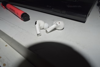 A pair of white wireless earbuds is lying on a white surface next to a red highlighter pen and a black laptop. There are some small black and red marks or damage on the earbuds. The scene is bathed in light, casting shadows along the surface.