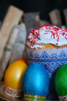 A close-up view of decorated Easter eggs with vibrant colors, including blue, yellow, and green. They are placed next to a dessert, likely a traditional Easter cake topped with white icing and colorful sprinkles. The cake has a decorative wrapper featuring blue geometric patterns.