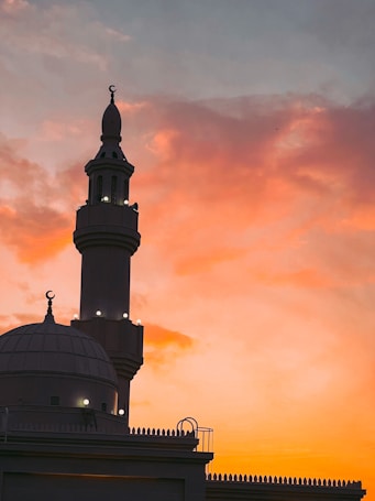 A mosque with a prominent minaret is silhouetted against a vibrant sunset sky. The sky displays a gradient of warm colors from orange to pink, creating a serene and captivating backdrop.