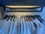 A row of neatly arranged white and light-colored garments hanging on black hangers in a closet. The hangers are placed on a metal rod, and the clothing includes various textures and patterns, primarily featuring lace and delicate fabrics. The area is softly illuminated, highlighting the details of the fabric.