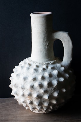 A textured ceramic jug with a bulbous, spiked surface and a narrow, cylindrical neck sits on a wooden surface. The light highlights its rough, matte finish against a dark background, accentuating its unique form and shadow.