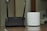 A black D-Link Wi-Fi router with two antennas is placed next to a white cylindrical Deco device on a wooden surface. The background appears to be a blurred indoor setting.