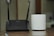 A black D-Link Wi-Fi router with two antennas is placed next to a white cylindrical Deco device on a wooden surface. The background appears to be a blurred indoor setting.