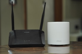 A black D-Link Wi-Fi router with two antennas is placed next to a white cylindrical Deco device on a wooden surface. The background appears to be a blurred indoor setting.