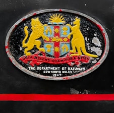 An oval-shaped emblem with a coat of arms featuring a lion and a kangaroo flanking a shield. The shield includes symbols under a rising sun, with a ribbon displaying Latin text beneath. Below the emblem, the text reads 'The Department of Railways, New South Wales, 1947'. The emblem is set against a dark background with a red horizontal stripe beneath it.