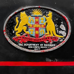 An oval-shaped emblem with a coat of arms featuring a lion and a kangaroo flanking a shield. The shield includes symbols under a rising sun, with a ribbon displaying Latin text beneath. Below the emblem, the text reads 'The Department of Railways, New South Wales, 1947'. The emblem is set against a dark background with a red horizontal stripe beneath it.