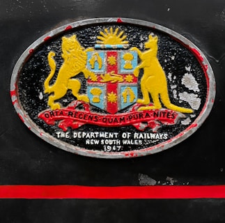 An oval-shaped emblem with a coat of arms featuring a lion and a kangaroo flanking a shield. The shield includes symbols under a rising sun, with a ribbon displaying Latin text beneath. Below the emblem, the text reads 'The Department of Railways, New South Wales, 1947'. The emblem is set against a dark background with a red horizontal stripe beneath it.