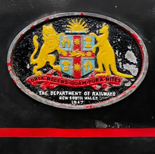 An oval-shaped emblem with a coat of arms featuring a lion and a kangaroo flanking a shield. The shield includes symbols under a rising sun, with a ribbon displaying Latin text beneath. Below the emblem, the text reads 'The Department of Railways, New South Wales, 1947'. The emblem is set against a dark background with a red horizontal stripe beneath it.